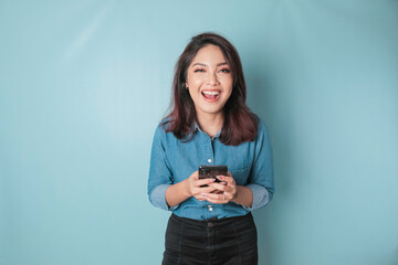 Wall Mural - A portrait of a happy Asian woman is smiling and holding her smartphone wearing a blue shirt isolated by a blue background