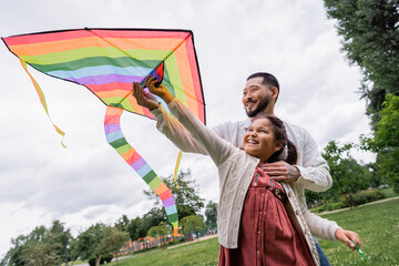 Positive asian dad hugging daughter with flying kite in summer park.