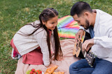 Fototapeta Londyn - Cheerful asian girl looking at father playing acoustic guitar near fruits and croissants on blanket in park.