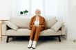 © Tatiana - a sweet, amiable elderly woman in a brown suit is sitting on a beige wide sofa, smiling pleasantly while in the comfortable environment of her apartment