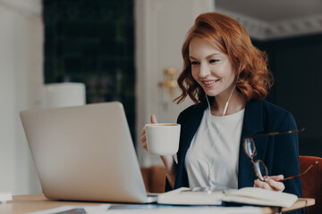 Young female teacher conducts lesson online, talks wih pupils via laptop computer, organizes video conference, teaches students, drinks aromatic coffee, holds glasses in hand, dressed formally