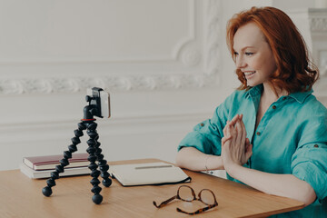 Cheerful ginger European woman sits in front of cellular camera, records self presentation video, shares professional skills, films educational lecture, poses at desktop, greets subscribers.