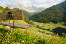 (Selective focus) Defocused park bench in the foreground with the Church of St. John (San Giovanni in Ranui) that stands out in the green meadows, in the heart of the beautiful Dolomitic landscape..