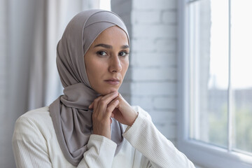 Wall Mural - Portrait of young beautiful Muslim woman near window at home, Arab woman in hijab looking at camera with concentration, close up photo