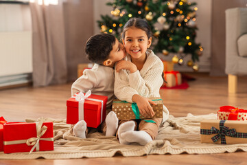 christmas, winter holidays and childhood concept - little boy kissing his happy sister sitting on floor with gifts at home