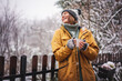 © olezzo - Young happy adult caucasian woman smiling in hat and yellow jacket enjoying tea in a mug with snowfall on the terrace of a country house