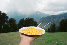 Close Up Shot Of Hand Holding Dish Of Noodles In Background Of Mountains In Manali, Himachal Pradesh, India. Tourist Enjoying Food In Mountains During Holiday Vacation.  