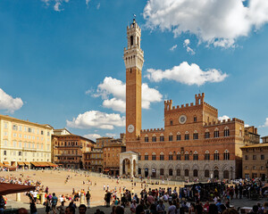 Tourists in the Piazza del Campo, the central square of the city of Sienna, Tuscany, Italy. Torre del Mangia tower rises behind