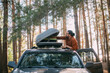 © Anna - A young man opens the upper tourist trunk of a car in a pine forest on the shore of a lake at sunset.