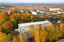 Aerial View Of School, College Or Kindergarten Building With Big Yard Among Autumn Trees On Rural Landscape Background