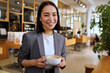 © insta_photos - Smiling young elegant professional leader Asian woman, female executive manager businesswoman, coffee house small business owner wearing suit holding coffee standing looking at camera, portrait.