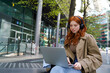 © insta_photos - Teen redhead hipster girl student wearing headphones using laptop computer tech device on city street online learning outdoors, elearning outside watching educational webinar sitting in urban park.