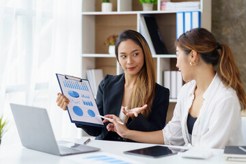 Wall Mural - Two professional young Asian business women discuss work, graph, chart using laptop computer for office work.