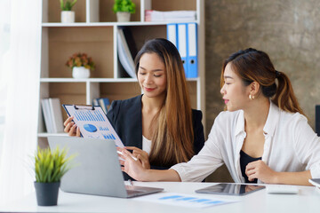 Wall Mural - Two professional young Asian business women discuss work, graph, chart using laptop computer for office work.