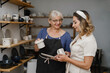 © ninelutsk - Potter artist showing her work in studio to customer, client. Mature ceramist standing at workshop with clay cups in hands. Successful lady potter own making porcelain products with student