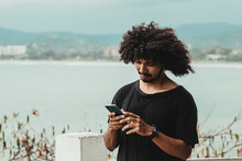 Black Man Chatting On Smartphone Against Sea In Town