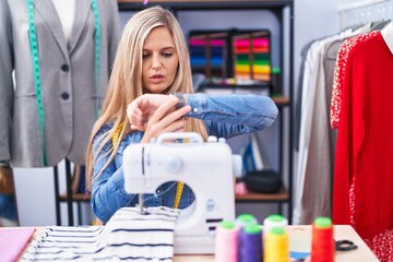 Sticker - Blonde woman dressmaker designer using sew machine checking the time on wrist watch, relaxed and confident