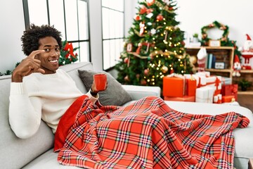 Sticker - Young african american man sitting on the sofa drinking coffee by christmas tree smiling and confident gesturing with hand doing small size sign with fingers looking and the camera. measure concept.