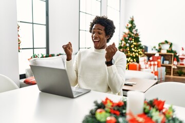 Poster - Young african american man using laptop sitting on the table by christmas tree celebrating surprised and amazed for success with arms raised and open eyes. winner concept.