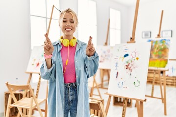 Canvas Print - Young caucasian girl at art studio gesturing finger crossed smiling with hope and eyes closed. luck and superstitious concept.