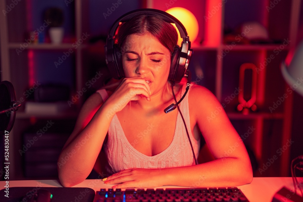 Young blonde woman playing video games wearing headphones looking stressed and nervous with hands on mouth biting nails. anxiety problem.