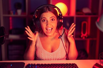 Poster - Young blonde woman playing video games wearing headphones celebrating crazy and amazed for success with arms raised and open eyes screaming excited. winner concept