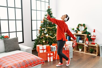 Sticker - Young hispanic woman listening to music dancing by christmas tree at home