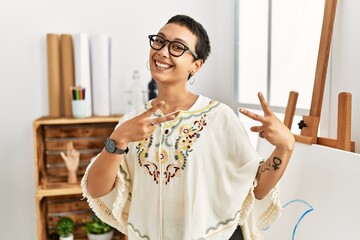 Poster - Young hispanic woman with short hair at art studio smiling looking to the camera showing fingers doing victory sign. number two.