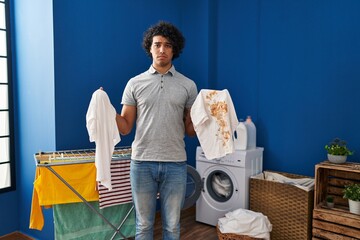 Sticker - Hispanic man with curly hair holding clean white t shirt and t shirt with dirty stain depressed and worry for distress, crying angry and afraid. sad expression.