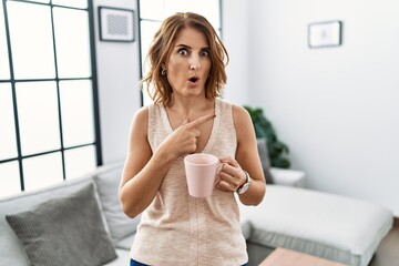 Poster - Middle age woman drinking a cup coffee at home surprised pointing with finger to the side, open mouth amazed expression.