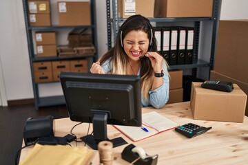 Wall Mural - Young hispanic woman working at small business ecommerce wearing headset screaming proud, celebrating victory and success very excited with raised arm