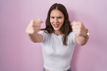 Poster - Young hispanic girl standing over pink background angry and mad raising fists frustrated and furious while shouting with anger. rage and aggressive concept.