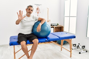 Canvas Print - Middle age hispanic man at pain recovery clinic holding pilates ball doing stop sing with palm of the hand. warning expression with negative and serious gesture on the face.