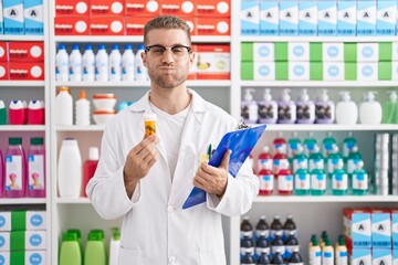 Sticker - Young caucasian man working at pharmacy drugstore holding pills puffing cheeks with funny face. mouth inflated with air, catching air.