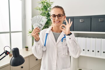 Canvas Print - Young doctor woman holding dollars banknotes doing ok sign with fingers, smiling friendly gesturing excellent symbol