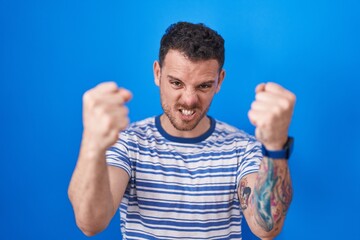 Poster - Young hispanic man standing over blue background angry and mad raising fists frustrated and furious while shouting with anger. rage and aggressive concept.