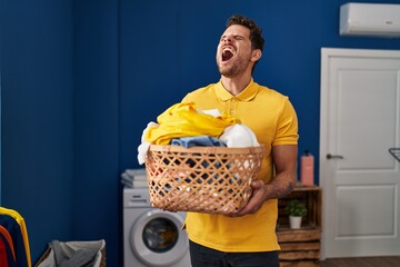 Poster - Young hispanic man holding laundry basket angry and mad screaming frustrated and furious, shouting with anger. rage and aggressive concept.