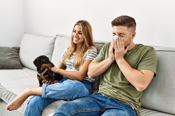 Wall Mural - Young hispanic couple sitting on the sofa with dog at home. Man using handkerchief.