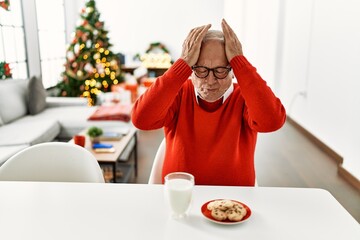 Canvas Print - Senior man with grey hair sitting on the table with cookies by christmas tree suffering from headache desperate and stressed because pain and migraine. hands on head.