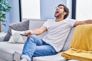 Wall Mural - Handsome latin man holding television remote control angry and mad screaming frustrated and furious, shouting with anger looking up.