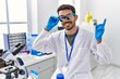 © Krakenimages.com - Young hispanic man working at scientist laboratory wearing magnifying glasses smiling happy pointing with hand and finger to the side