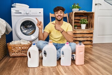 Wall Mural - Arab man with beard doing laundry sitting on the floor with detergent bottle smiling and looking at the camera pointing with two hands and fingers to the side.