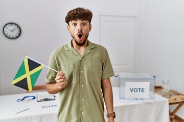 Canvas Print - Young arab man at political campaign election holding jamaica flag scared and amazed with open mouth for surprise, disbelief face