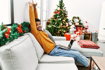 Canvas Print - Young arab man stretching arms using laptop sitting on the sofa by christmas tree at home.