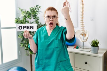 Poster - Young redhead physiotherapist woman working at pain recovery clinic holding open banner annoyed and frustrated shouting with anger, yelling crazy with anger and hand raised