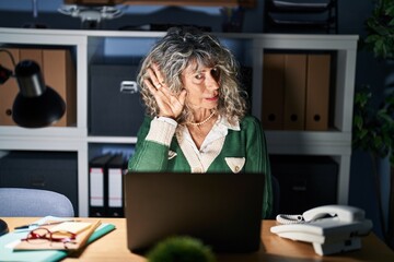 Wall Mural - Middle age woman working at night using computer laptop smiling with hand over ear listening an hearing to rumor or gossip. deafness concept.