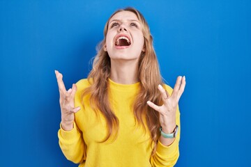 Wall Mural - Young caucasian woman standing over blue background crazy and mad shouting and yelling with aggressive expression and arms raised. frustration concept.