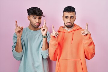 Wall Mural - Young hispanic gay couple standing over pink background pointing up looking sad and upset, indicating direction with fingers, unhappy and depressed.