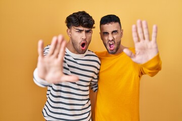 Wall Mural - Young hispanic gay couple standing over yellow background doing stop gesture with hands palms, angry and frustration expression