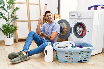 Wall Mural - Young hispanic man putting dirty laundry into washing machine smiling with happy face winking at the camera doing victory sign with fingers. number two.